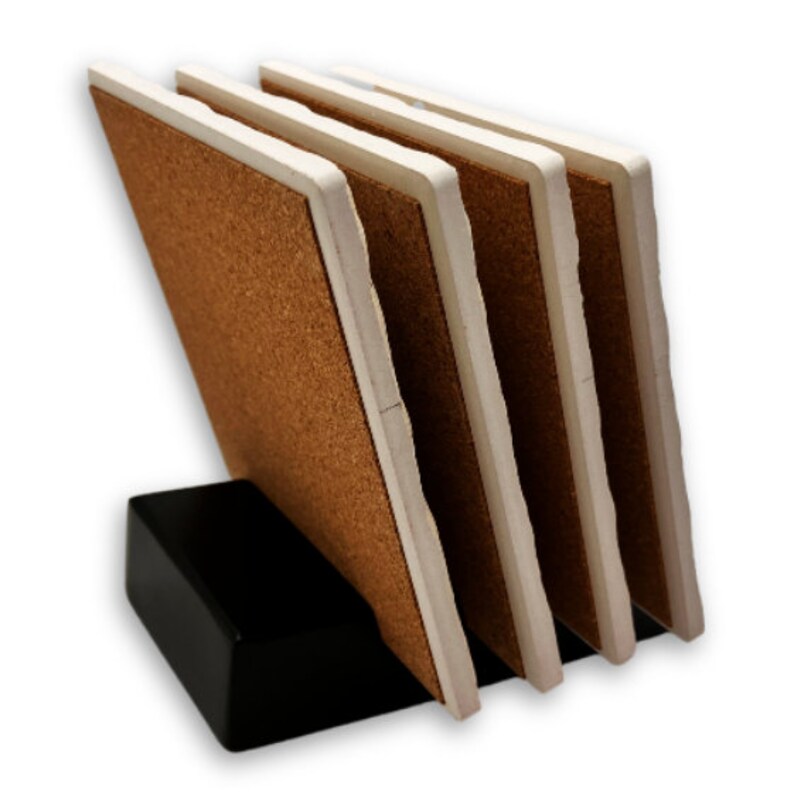 May include: A set of four square coasters with a cork surface and white edges, displayed in a black holder. The coasters are angled, showcasing their brown and white color scheme. The holder is a solid black rectangle.