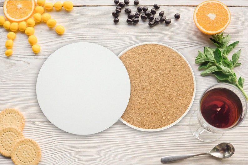 May include: Two round coasters, one white and one cork, on a white wooden surface. The white coaster is blank and the cork coaster has a natural brown colour. There are other items on the surface, including a bowl of yellow sweets, a bowl of chocolate sweets, a glass of tea, and a spoon.