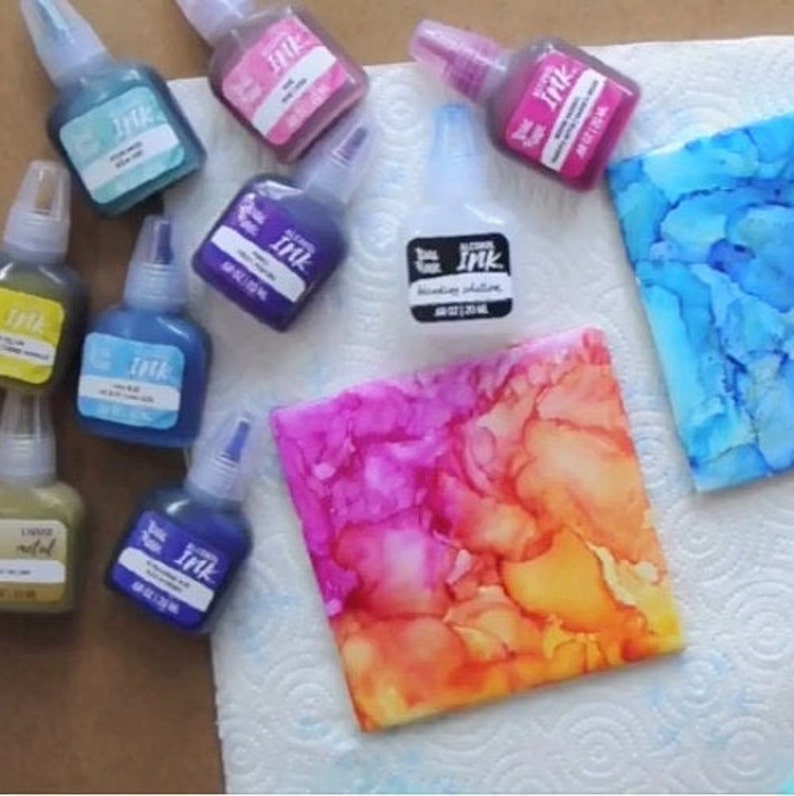 May include: A variety of alcohol inks in small bottles with dropper tops. The inks are in colors like blue, purple, pink, gold, and black. There are two pieces of paper with alcohol ink art on them. One is a blue and white abstract design and the other is a pink, orange, and yellow abstract design.