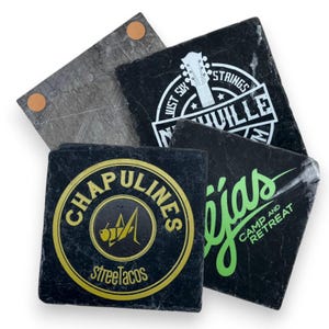 May include: A collection of square slate coasters. One coaster is gray, while the others are black with various white and gold logos and text. The coasters feature the text "Just Six Strings Nashville", "Chapulines street tacos", and "Tejas Camp and Retreat".