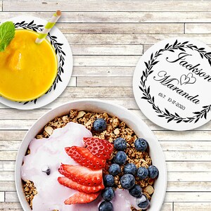 May include: Two white round coasters with black floral wreaths and the text "Jackson Maranda est. 10-17-2020" in a cursive font. The coasters are on a light wood surface with a bowl of granola, yogurt, strawberries, and blueberries.