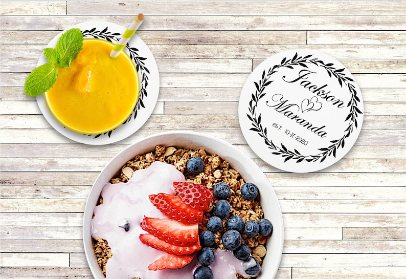May include: Two white round coasters with black floral wreaths and the text "Jackson Maranda est. 10-17-2020" in a cursive font. The coasters are on a light wood surface with a bowl of granola, yogurt, strawberries, and blueberries.