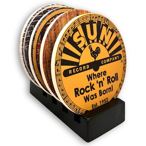May include: A set of round coasters with a black holder. The coasters feature various designs, including a vintage-style Sun Record Company logo with the text "Where Rock 'n' Roll Was Born!" and a rooster graphic. The holder is black.