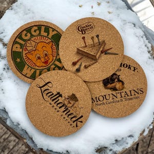 May include: Four cork coasters with different designs. The first coaster features a cartoon pig with the text "Piggly Wiggly". The second coaster features a wooden triangle with golf tees and the text "Cracker Barrel". The third coaster features a cartoon woodcarver with the text "Leatherneck Woodcarver". The fourth coaster features a black bear with the text "Smoky Mountains North Carolina & Tennessee".