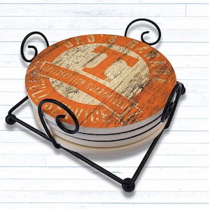 May include: Set of four orange and white coasters with a black metal holder. The coasters feature a distressed, vintage design with the text "Dad's Garage" and a large orange letter "T".