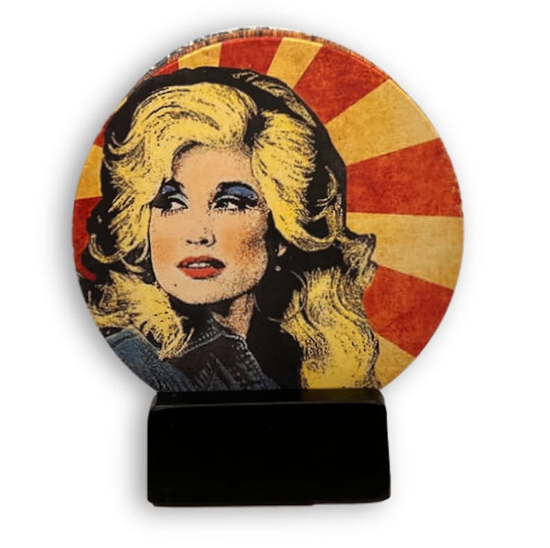 May include: A pop art style image of a woman with blonde hair, red lips, and blue eyeshadow, set against a red and yellow sunburst background. The image is mounted on a black rectangular base, creating a decorative item.