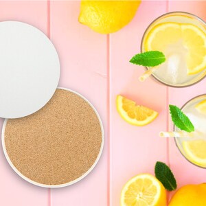 May include: Two white coasters, one with a cork surface and the other blank, on a pink background with lemons and glasses of lemonade.