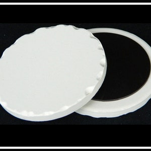 May include: A white ceramic round magnet with a scalloped edge. The magnet is in two parts, one with a white surface and the other with a black surface.