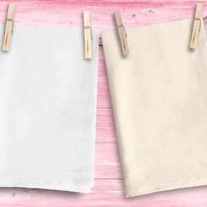 May include: Two white fabric tea towels hanging on a pink wooden background. The tea towels are hanging from clothespins. One tea towel is white and the other is a cream color.
