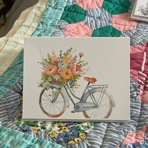 May include: A greeting card featuring a watercolor illustration of a vintage bicycle carrying a basket overflowing with colorful flowers. The bicycle is light blue with orange accents, set against a white background.
