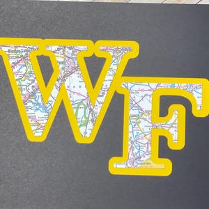 May include: A black rectangular sign featuring the letters WF in a bold, block font. The letters are outlined in bright yellow and filled with a colorful road map pattern showing various highways, city names, and geographic details in blue, red, and green.