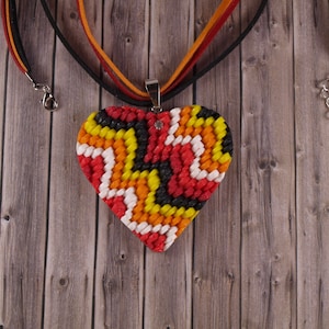 Peut inclure: Un collier avec un pendentif en forme de cœur avec un motif géométrique coloré en rouge, orange, jaune, noir et blanc. Le collier est fait d'un matériau tissé et a un fermoir argenté.