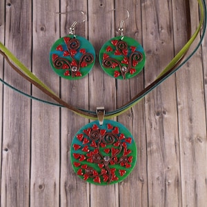 May include: A set of handmade jewellery featuring a necklace and earrings with a tree design. The tree is made of red hearts and brown branches on a green and blue background.