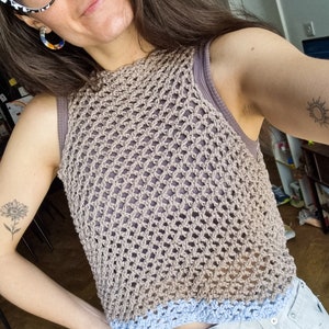 May include: A brown crochet top with a blue trim. The top is sleeveless and has a loose fit. The model is wearing the top with a pair of light blue jeans.