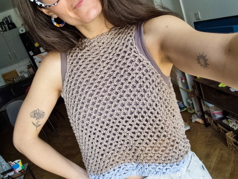 May include: A brown crochet top with a blue trim. The top is sleeveless and has a loose fit. The model is wearing the top with a pair of light blue jeans.