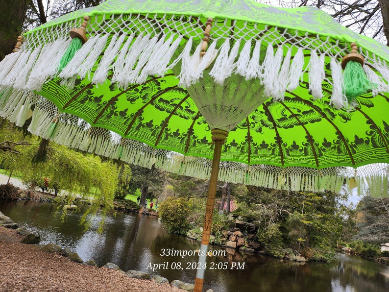 Pu&ograve; includere: Un ombrello con motivo verde e bianco con bordi a frangia pende sopra uno stagno. L'ombrello ha un palo di legno e un fiocco verde e bianco. Il testo "33imports.com April 08, 2024 2:05 PM" &egrave; visibile nell'angolo in basso a destra dell'immagine.