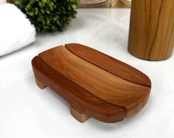 May include: A wooden soap dish with a rectangular shape and a slatted design. The dish is made of a warm, reddish-brown wood and has small feet. The soap dish is designed to hold a bar of soap and allow for drainage.