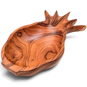 May include: A wooden bowl carved into the shape of a pineapple, featuring a smooth, polished surface with prominent brown and tan wood grain patterns. The bowl has a wide, rounded base and a pointed, leafy top section carved from the wood.
