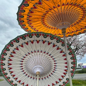 May include: Two colorful, hand-painted paper umbrellas with intricate designs. The top umbrella is yellow with red and blue accents, and the bottom umbrella is white with red, blue, and green accents.