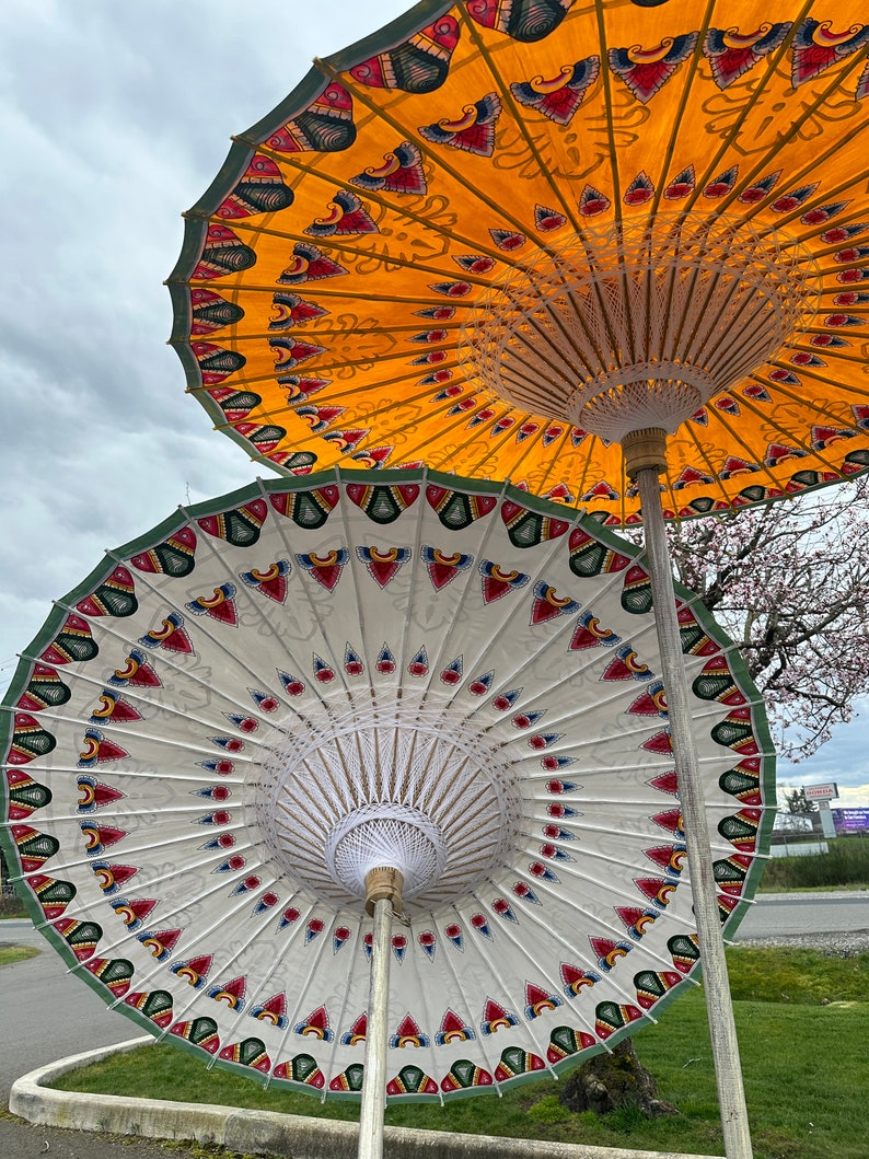 May include: Two colorful, hand-painted paper umbrellas with intricate designs. The top umbrella is yellow with red and blue accents, and the bottom umbrella is white with red, blue, and green accents.