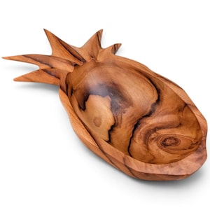 May include: A carved wooden bowl shaped like a pineapple, featuring a natural light and dark brown wood grain pattern. The bowl has a smooth, polished interior and pointed, leaf-like extensions at one end, mimicking the top of a pineapple.