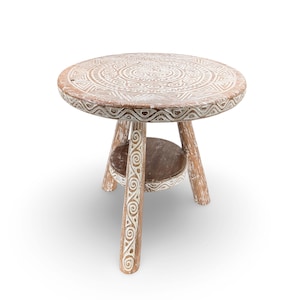 May include: A small, round wooden side table with a white, carved geometric design on the tabletop. The table has three legs and a lower shelf. The wood has a natural, distressed finish, with white accents.