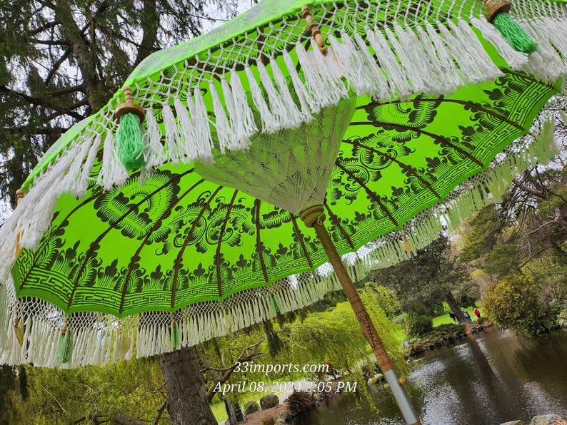 Pu&ograve; includere: Un ombrello verde e bianco con frange bianche e nappe verdi. L'ombrello &egrave; aperto e la parte superiore &egrave; visibile. L'ombrello &egrave; in un giardino con alberi e uno stagno sullo sfondo.
