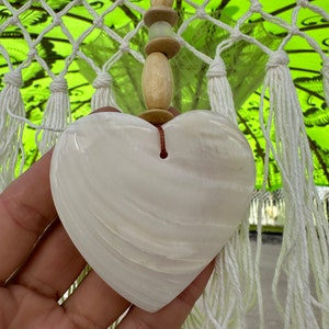 Pu&ograve; includere: Un ornamento a forma di cuore in conchiglia bianca con uno spago marrone appeso in cima. La conchiglia ha una superficie liscia e lucida e una delicata texture ondulata.