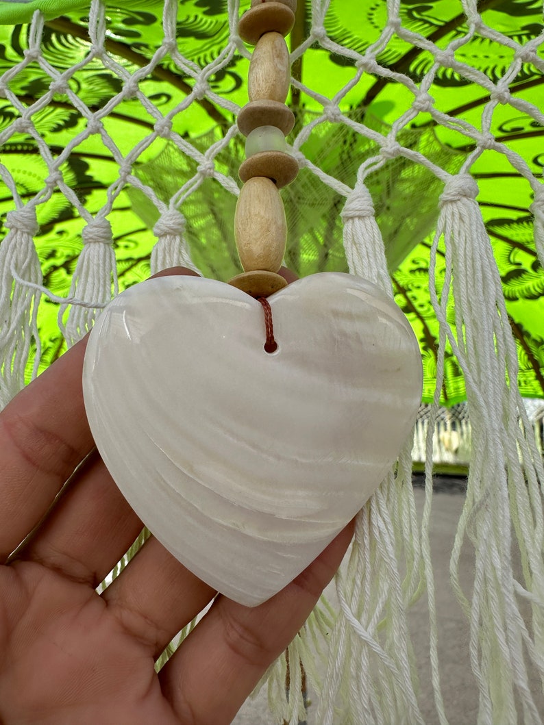 Pu&ograve; includere: Un ornamento a forma di cuore in conchiglia bianca con uno spago marrone appeso in cima. La conchiglia ha una superficie liscia e lucida e una delicata texture ondulata.