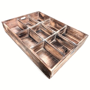 May include: A rectangular wooden tray with a distressed brown finish, featuring eight individual compartments of varying sizes. The tray includes integrated cutout handles on the shorter sides and exhibits a visible wood grain texture throughout.