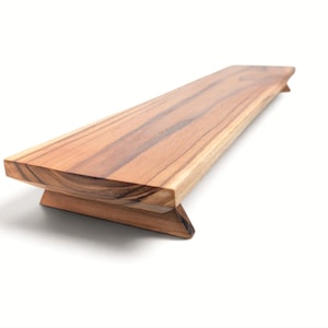 May include: A rectangular wooden riser with a natural light and dark brown grain pattern. The platform features a smooth, flat top surface supported by two angled, wedge-shaped feet at each end, elevating the board slightly above the surface.
