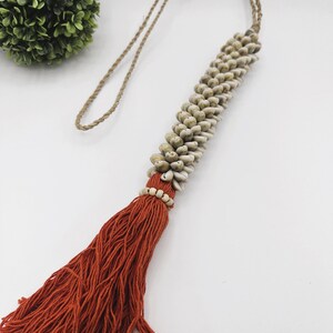 May include: A long, orange tassel with a string of white seashells and white beads at the top. The tassel is attached to a natural twine cord.