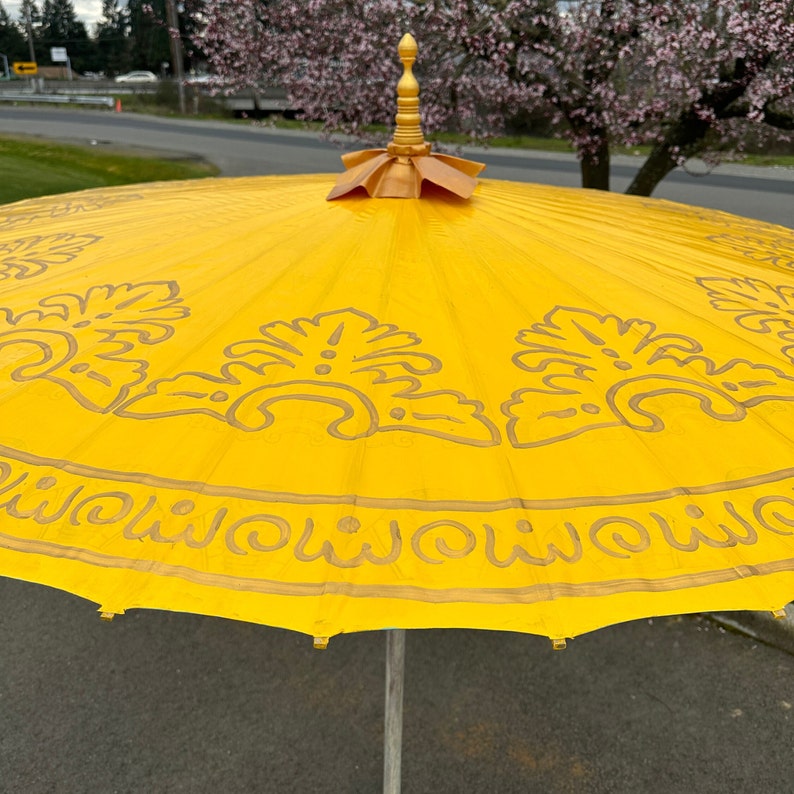 May include: A yellow umbrella with a gold trim and intricate floral designs. The umbrella has a wooden handle with a gold finial.