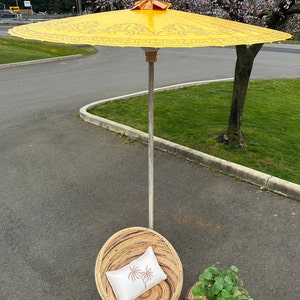 May include: A yellow and gold patterned umbrella with a wooden handle stands over a wicker chair with a white pillow and a wicker side table with a green plant.