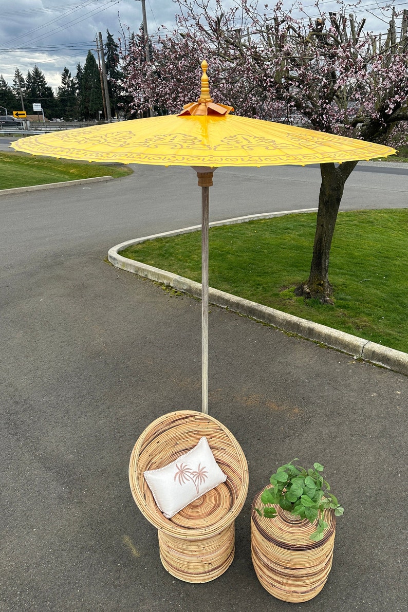 May include: A yellow and gold patterned umbrella with a wooden handle stands over a wicker chair with a white pillow and a wicker side table with a green plant.