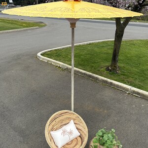 May include: A yellow and white patterned umbrella with a wooden handle, a wicker chair with a white pillow, and a wicker side table with a green plant.