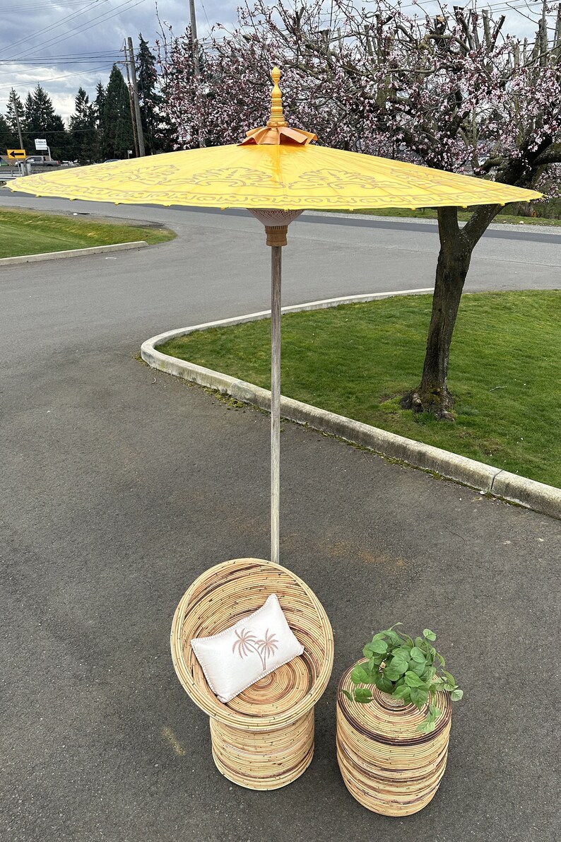 May include: A yellow and white patterned umbrella with a wooden handle, a wicker chair with a white pillow, and a wicker side table with a green plant.