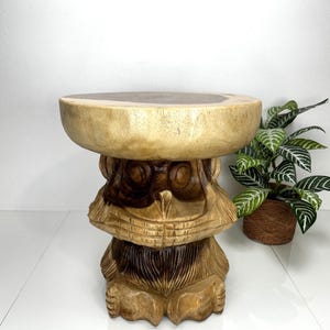 May include: A wooden stool carved in the shape of a face with large eyes. The stool is made of light brown wood with darker brown accents. The top of the stool is a round, flat surface.