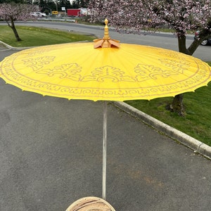 May include: A yellow and gold umbrella with a decorative pattern. The umbrella is open and the pole is visible. The umbrella is on a paved surface.