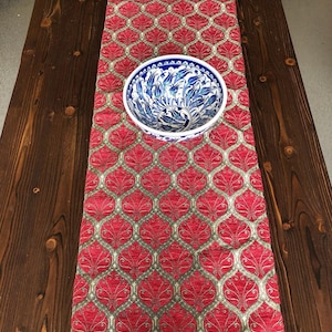 May include: A red table runner with a gold and white geometric pattern. The runner has a gold fringe border and is placed on a dark wood table. A blue and white ceramic bowl is centered on the runner.