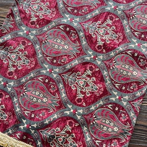 Può includere: Un runner da tavolo rosso e verde con un motivo paisley e frange dorate. Il runner ha un bordo a frange.