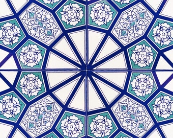 Op de afbeelding: Een witte tegel met een blauw en groen geometrisch patroon. Het patroon heeft een herhalend ontwerp van bloemen en ingewikkelde vormen.