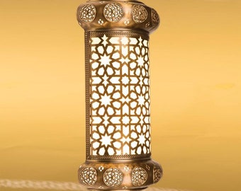 May include: Ornate bronze-colored pendant lamp with intricate star-shaped cutouts. The lamp has a cylindrical body with decorative circular accents at the top and bottom. The lamp is suspended against a warm, golden background.