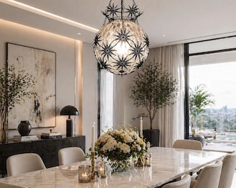 May include: A decorative chandelier with a globe-shaped glass shade adorned with black starburst designs hangs above a marble dining table. The dining room features a modern aesthetic with neutral tones, a large painting, and a floral centerpiece.