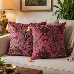 Op de afbeelding: Twee decoratieve kussens met een bloemenpatroon in roze, groene en rode tinten. Elk kussen heeft een gouden kwast. De kussens liggen op een beige bank.