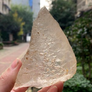 Può includere: Un grande cristallo di quarzo traslucido con una superficie ruvida e strutturata. Il cristallo è tenuto in mano, mostrando la sua forma naturale e irregolare e le inclusioni marrone chiaro. I bordi del cristallo sono frastagliati e il colore generale è un bianco lattiginoso pallido.