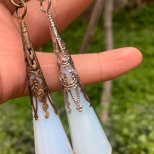 Puede incluir: Dos colgantes de lágrima de ópalo blanco iridiscente con engastes de filigrana plateada y bronce.