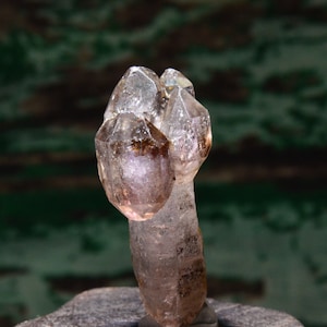 May include: A clear quartz crystal with a pink hue and brown inclusions. The crystal is shaped like a cluster of points and is sitting on a wooden base.
