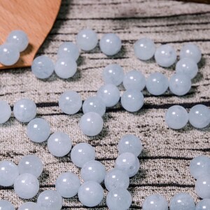 May include: A collection of light blue round beads made of aquamarine. The beads are scattered on a textured fabric surface.