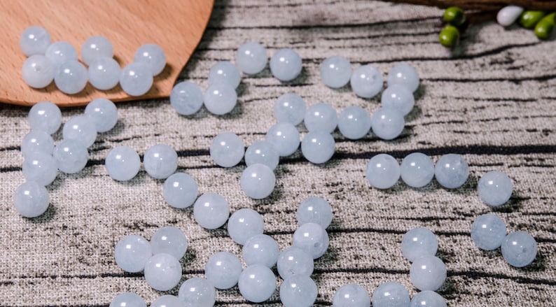 May include: A collection of light blue round beads made of aquamarine. The beads are scattered on a textured fabric surface.